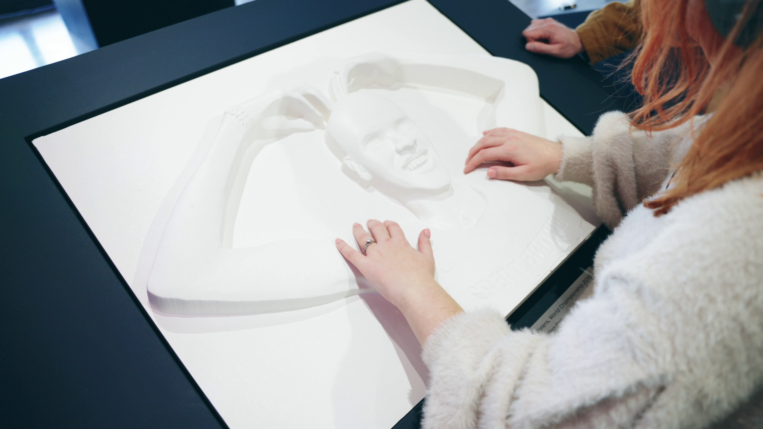 vlcsnap-2025-09-19-15h16m49s310 A young lady feels a tactile carving of Mo Farah and understands what he looks like for the first time. Mo Farah is standing in his classic 'Mobot' pose he made famous, with his arms in an 'M' shape above his head.